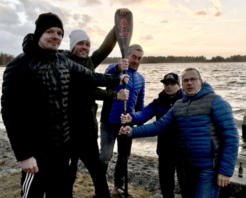 5 Paddler stehen an einem See und halten gemeinsam ein Paddel.