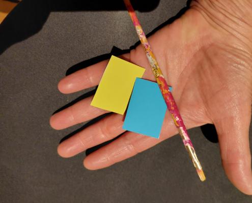 Auf einer Hand liegen eine gelbe und blaue kleine Farbkarte sowie ein Pinsel.
