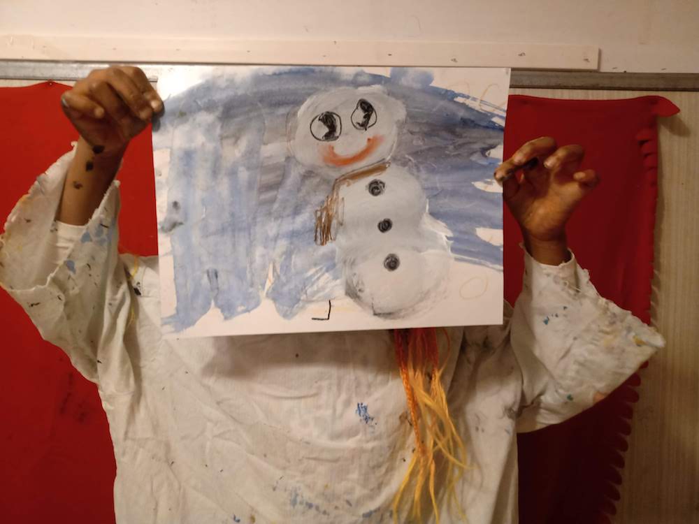 Kind hält ein Bild vor einem Gesicht hoch. Es zeigt einen Schneemann.